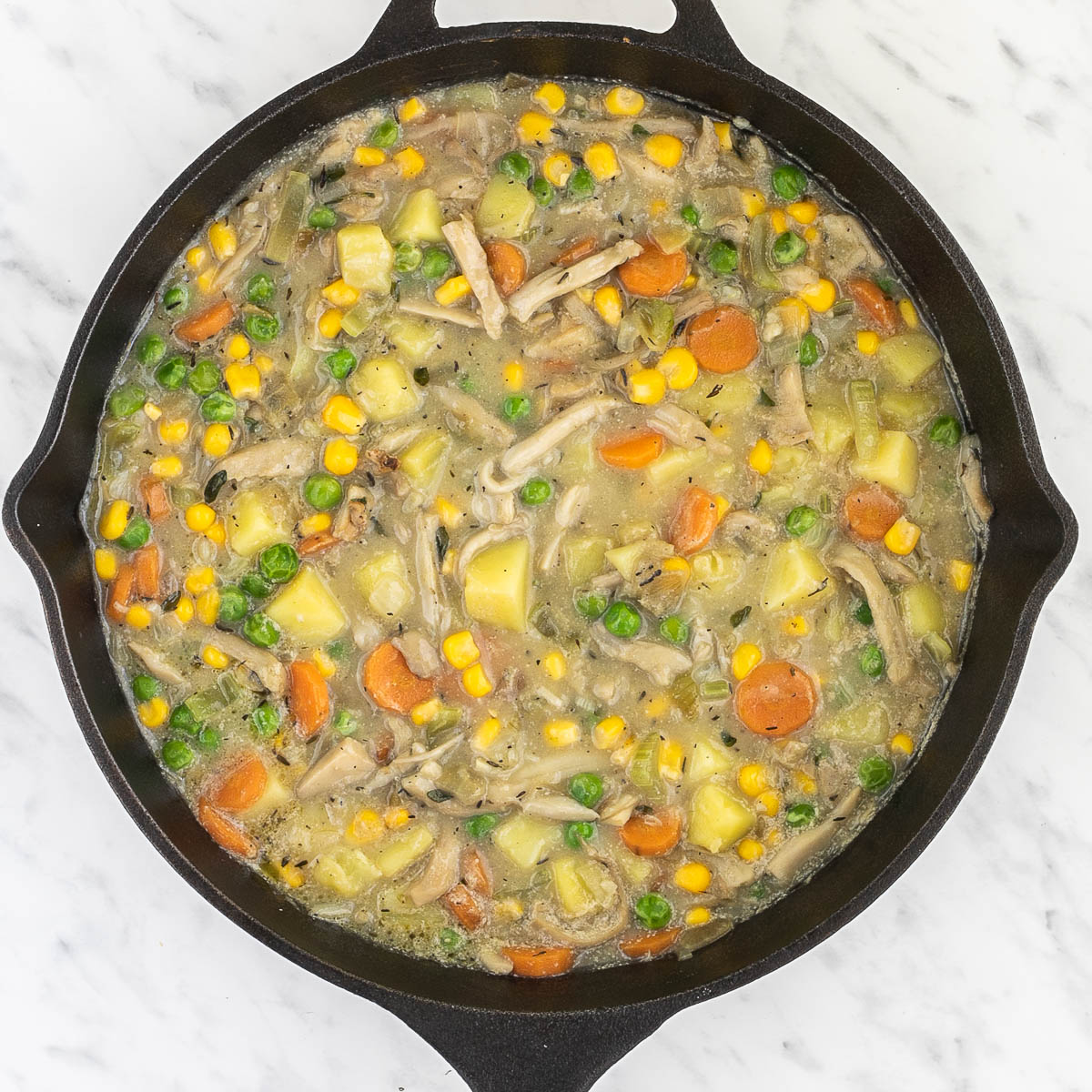 Cast iron skillet with light brown thick stew full of sliced carrots, chopped onion, garlic, celery, potatoes, corn, peas, shredded king oyster mushrooms, bay leaves, thyme.