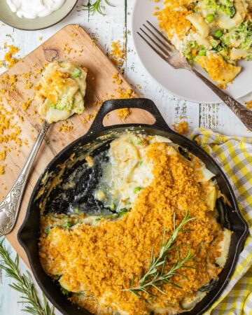 A black cast iron skillet with potato slices in a yellow thick sauce topped with yellow breadcrumbs. A part of it is missing and there is a gray plate with some food on it where you can see peas and broccoli as well.