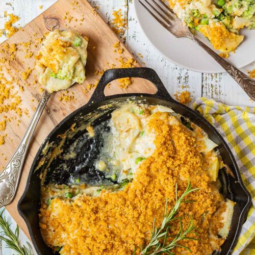 A black cast iron skillet with potato slices in a yellow thick sauce topped with yellow breadcrumbs. A part of it is missing and there is a gray plate with some food on it where you can see peas and broccoli as well.
