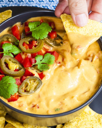 Yellow sauce in a black bowl topped with red veggie chunks, sliced green pepper and green leaves of herbs. A tortilla chips is dipped in it. Other tortilla chips are scattered around.