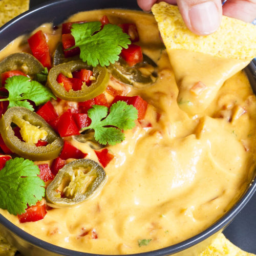 Yellow sauce in a black bowl topped with red veggie chunks, sliced green pepper and green leaves of herbs. A tortilla chips is dipped in it. Other tortilla chips are scattered around.