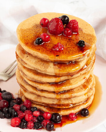 Pink plate with a stack of pancakes topped with black and red berries and maple syrup