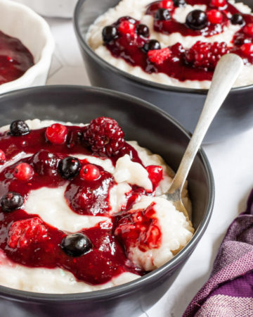A black bowl up close with white pudding topped with a swirl of thick red-purple sauce and different berries. A spoon is taking the first bite. Another bowl is in the background.