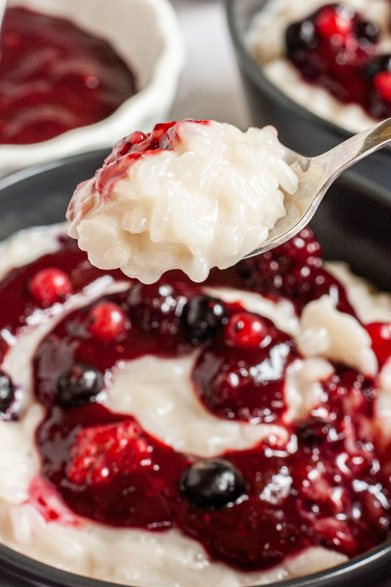 A spoon of creamy white rice pudding. In the background a black bowl with white pudding topped with a swirl of thick red-purple sauce and different berries.