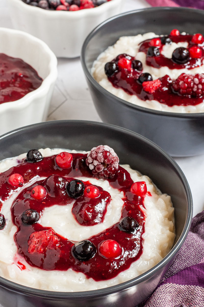 A black bowl up close with white pudding topped with a swirl of thick red-purple sauce and different berries. Another bowl is in the background.