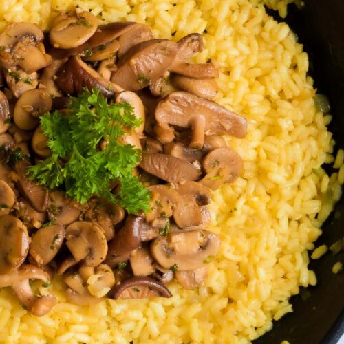 Large black frying pan with yellow creamy rice topped with brown mushroom slices and parsley leaves.