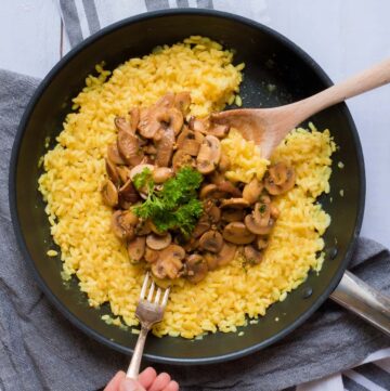 Large black frying pan with yellow creamy rice topped with brown mushroom slices and parsley leaves. A hand is holding a fork and taking some. A wooden spatula is placed in it as well.