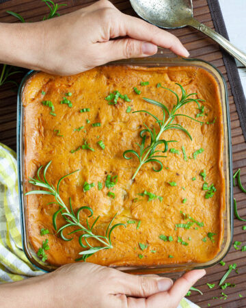A glass casserole dish with layers of potato slices with an orange creamy sauce sprinkled with chopped parsley and rosemary. two hand is holding the sides.
