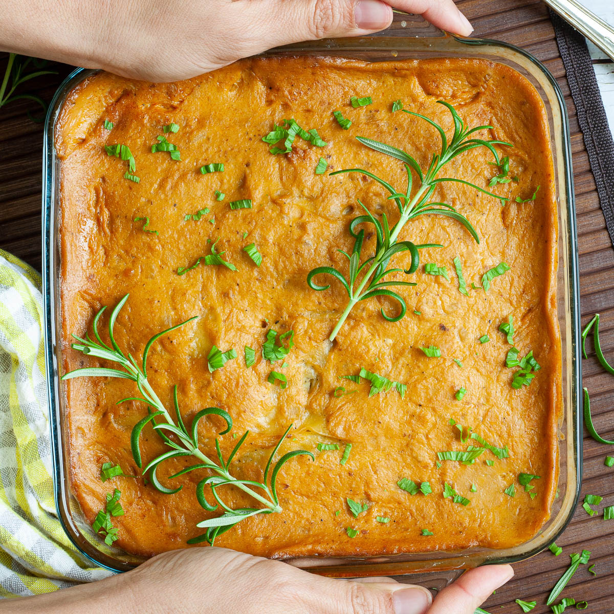 A glass casserole dish with layers of potato slices with an orange creamy sauce sprinkled with chopped parsley and rosemary. Two hand is holding the sides.