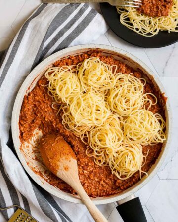 White frying pan with red thick sauce with fake ground meat pieces topped with small heaps of spaghetti. A wooden spatula is placed inside. A small black plate with a serving serving is placed next to it.