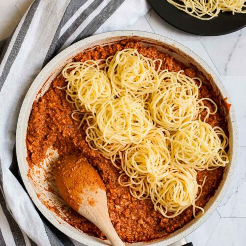 White frying pan with red thick sauce with fake ground meat pieces topped with small heaps of spaghetti. A wooden spatula is placed inside. A small black plate with a serving serving is placed next to it.