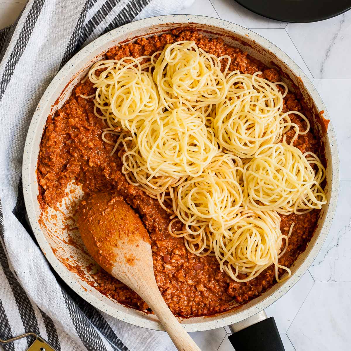 White frying pan with red thick sauce with fake ground meat pieces topped with small heaps of spaghetti. A wooden spatula is placed inside.