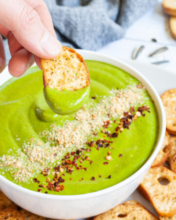 A white bowl with green sauce sprinkled with yellow flakes and red chili flakes. A hand is holding a piece of toasted bread to dip it in. More small toasted bread pieces are around the bowl.