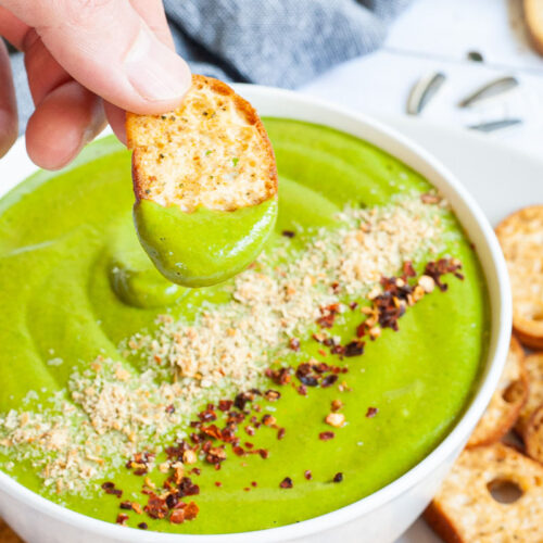 A white bowl with green sauce sprinkled with yellow flakes and red chili flakes. A hand is holding a piece of toasted bread to dip it in. More small toasted bread pieces are around the bowl.
