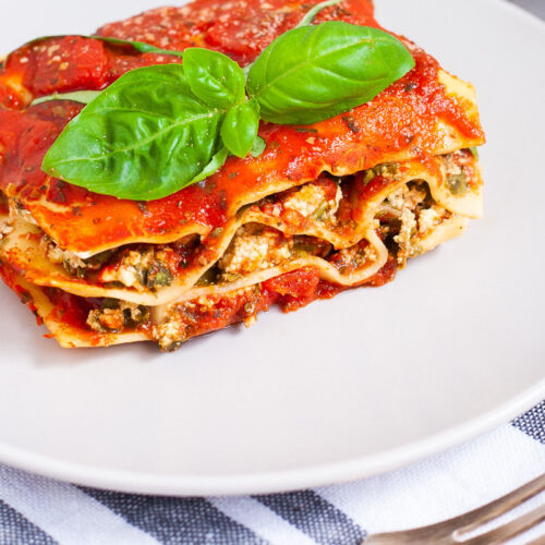 A white plate with a slice of lasagna where you can see the layers of red sauce, pasta sheets and a white ricotto and chopped spinach leaves.