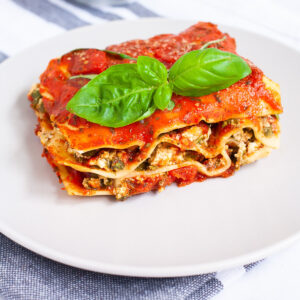 A white plate with a slice of lasagna where you can see the layers of red sauce, pasta sheets and a white ricotto and chopped spinach leaves.