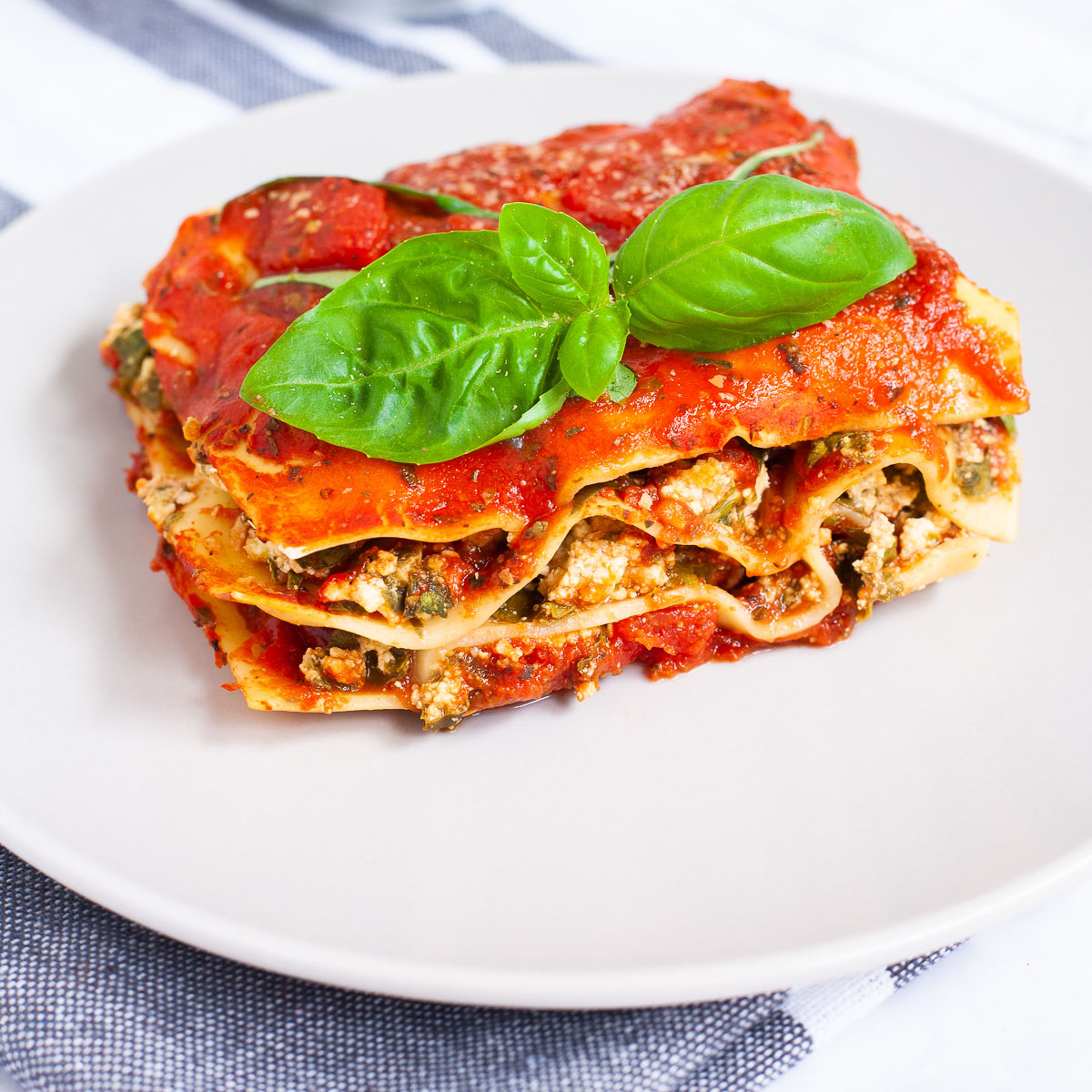 A white plate with a slice of lasagna where you can see the layers of red sauce, pasta sheets and a white ricotto and chopped spinach leaves.