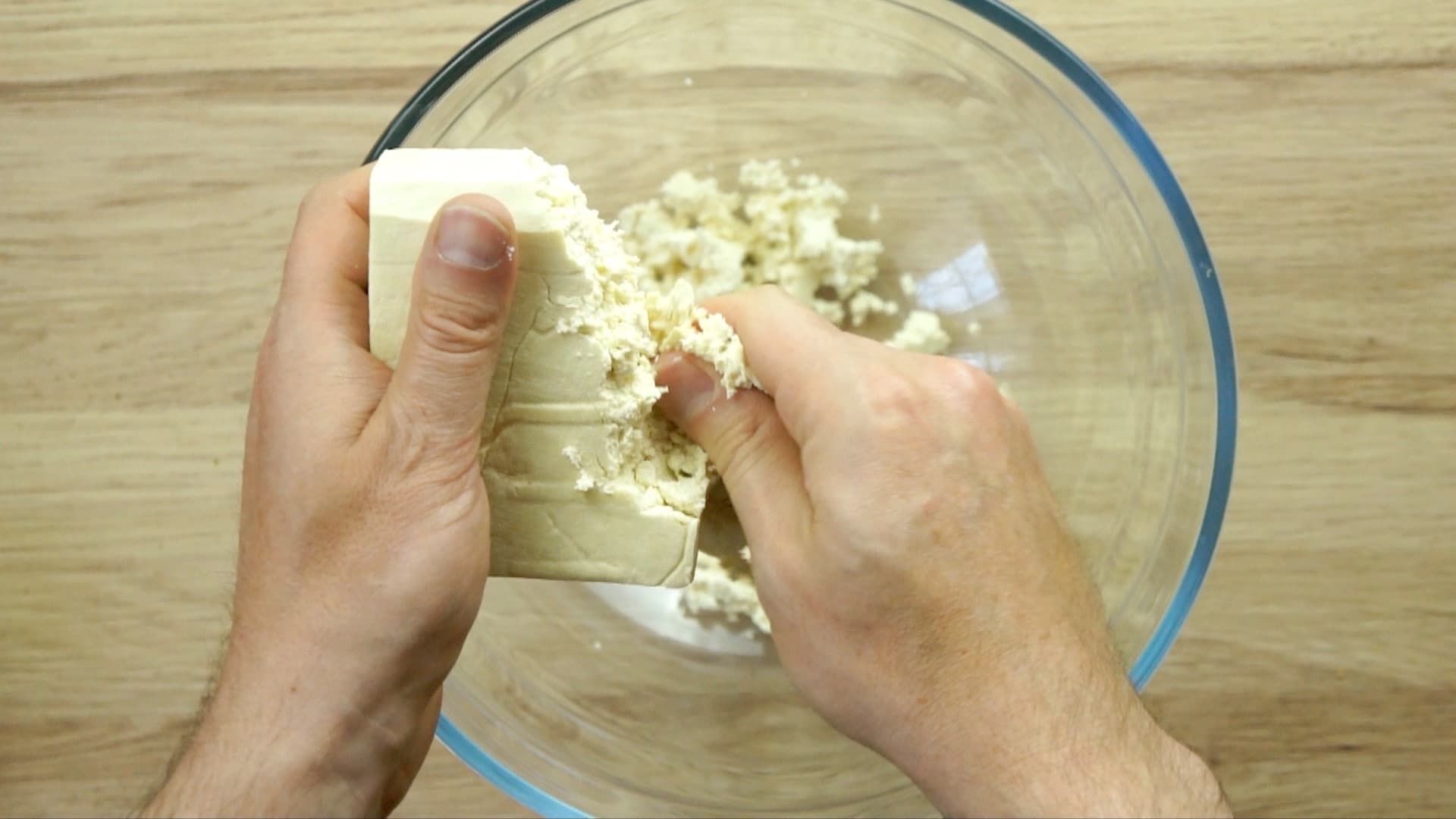 A hand is crumbling a block of tofu into a glass mixing bowl.