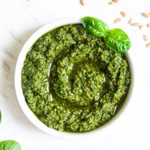 Small white bowl with a chunky green paste and a fresh basil leaf on the side. Fresh spinach leaves and sunflower seeds are scattered around.