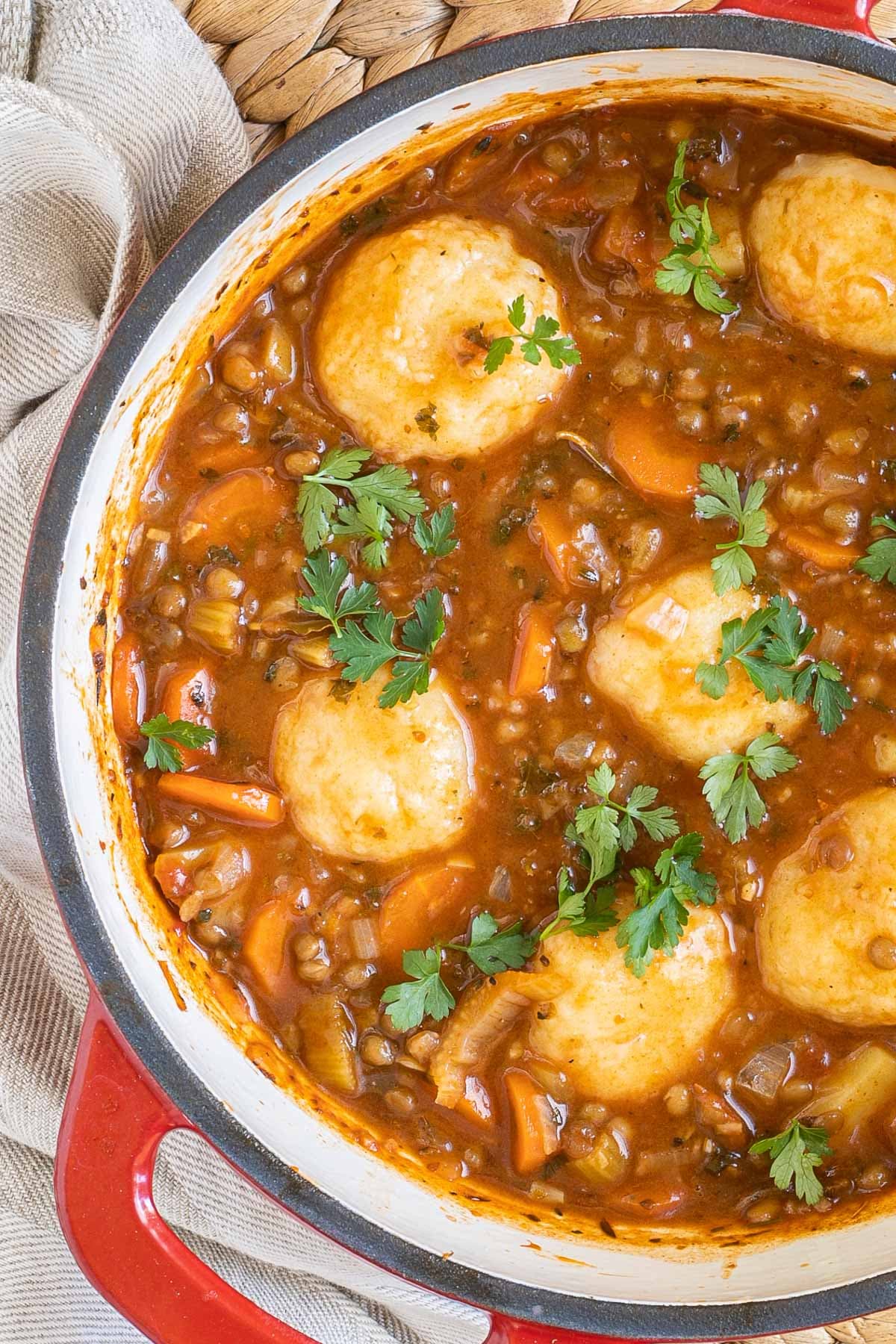 A red-white Dutch oven with thick brown stew, lentils, chopped veggies and a couple of dumplings.