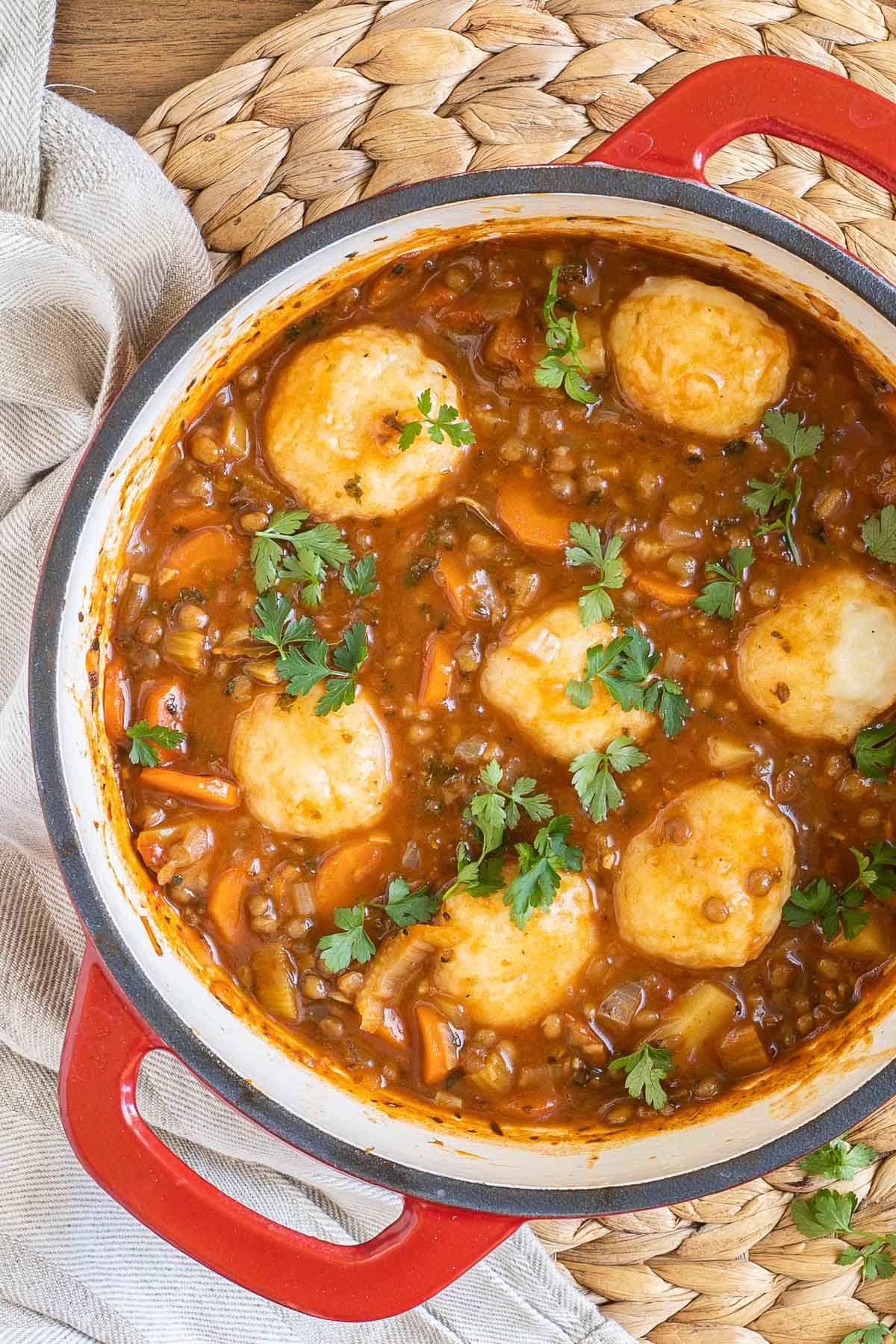 A red-white Dutch oven with thick brown stew, lentils, chopped veggies and a couple of dumplings.