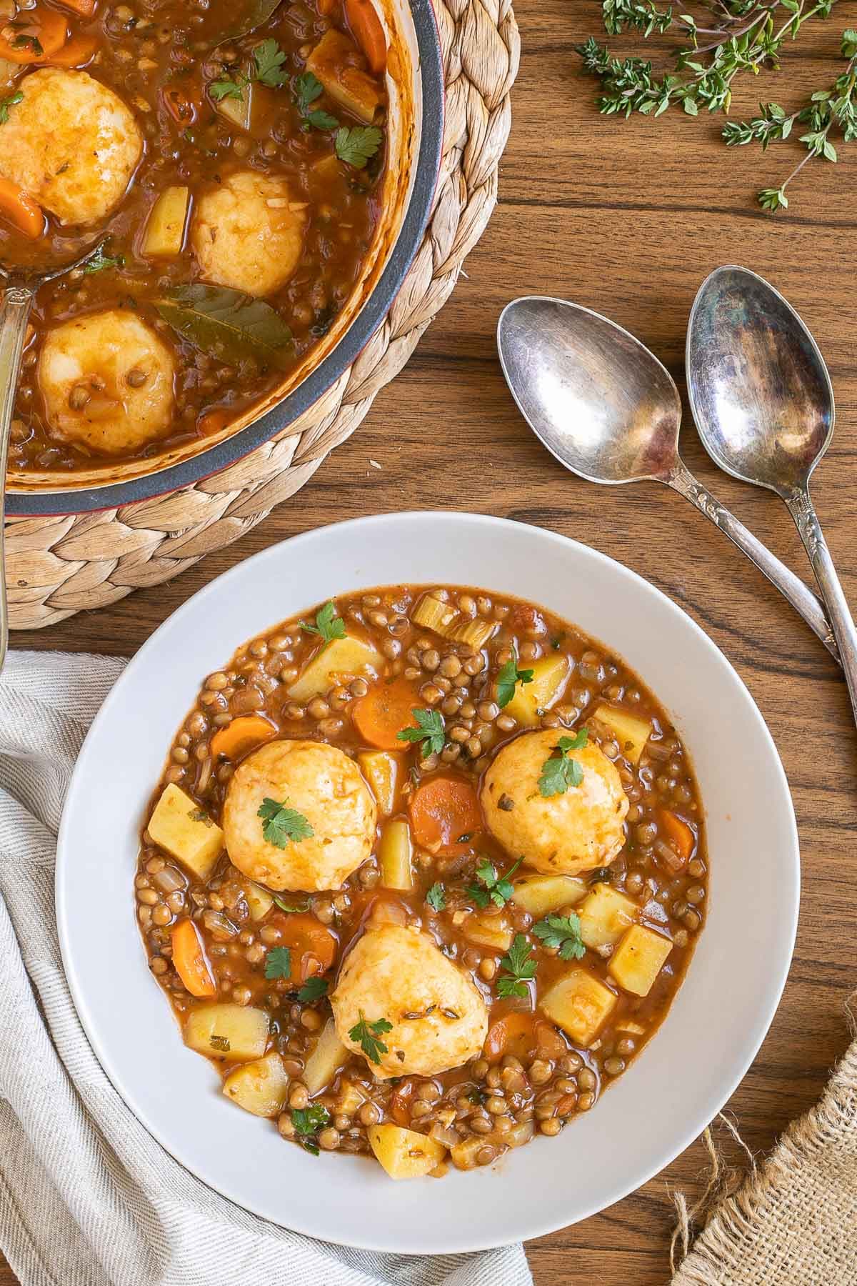 A white bowl with thick brown stew, lentils, chopped veggies and a couple of dumplings. The remaining stew is in a red-white Dutch oven.