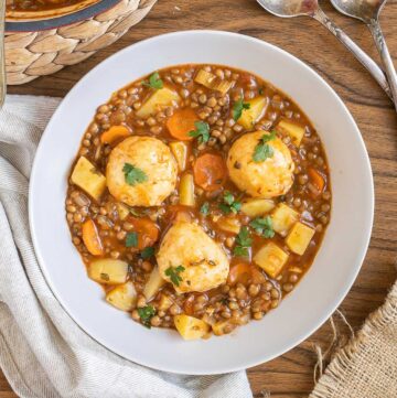 A white bowl with thick brown stew, lentils, chopped veggies and a couple of dumplings. The remaining stew is in a red-white Dutch oven.