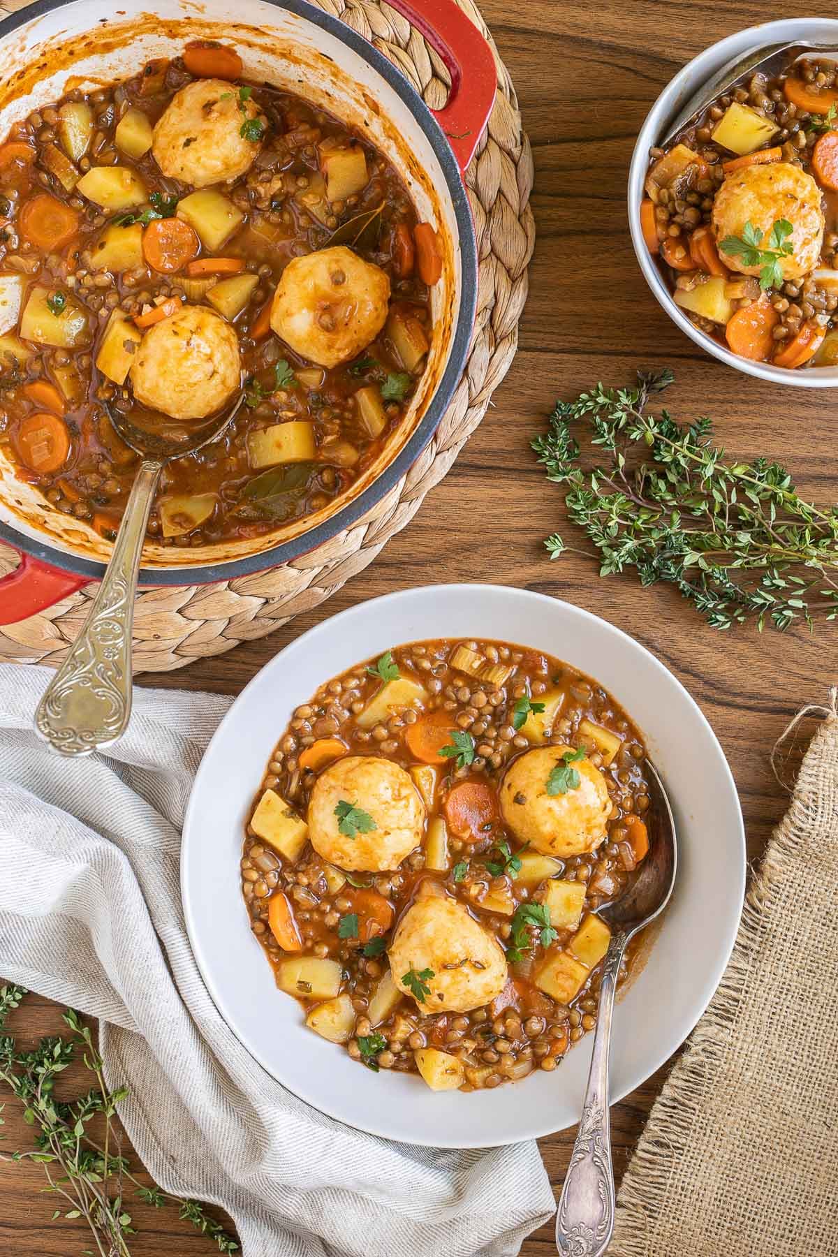 2 white bowls with thick brown stew, lentils, chopped veggies and a couple of dumplings. The remaining stew is in a red-white Dutch oven.