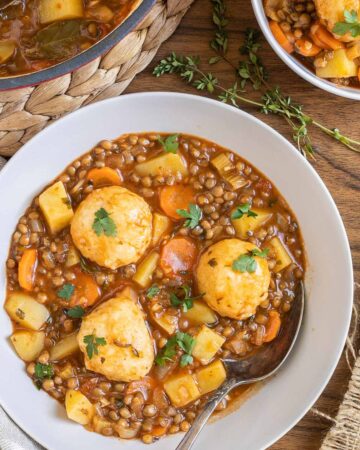 2 white bowls with thick brown stew, lentils, chopped veggies and a couple of dumplings. The remaining stew is in a red-white Dutch oven.