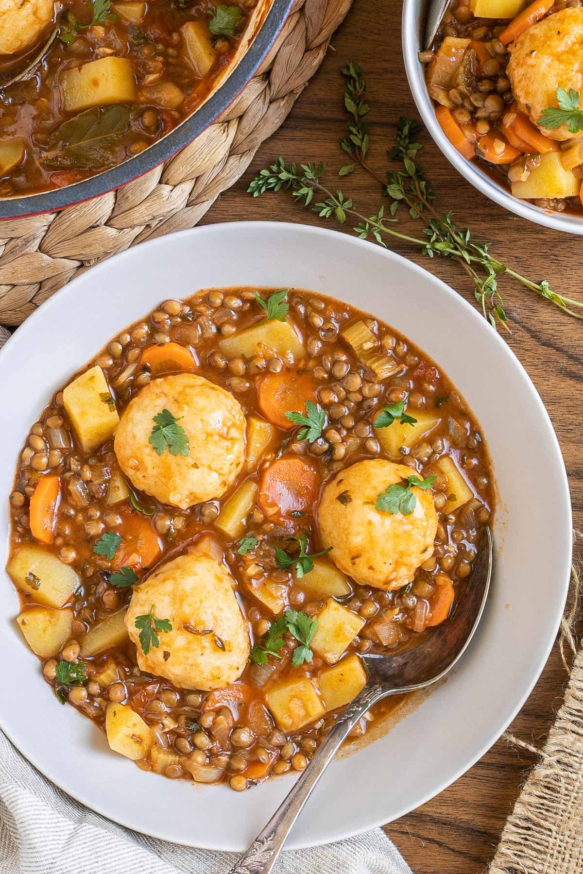 2 white bowls with thick brown stew, lentils, chopped veggies and a couple of dumplings. The remaining stew is in a red-white Dutch oven.