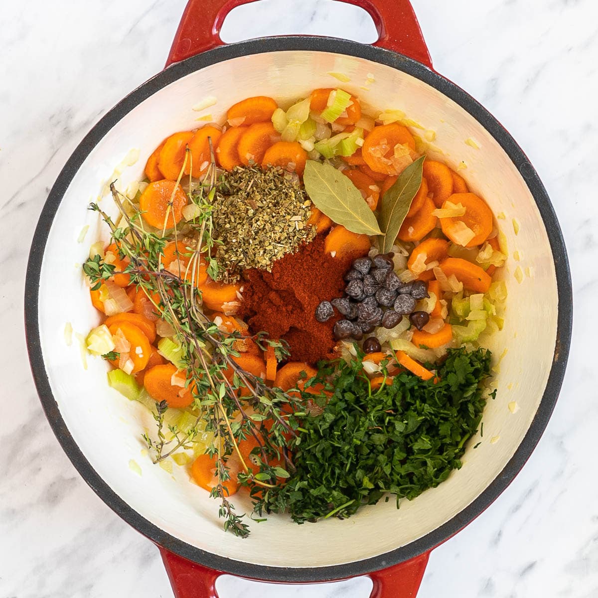 A red-white Dutch oven with chopped onion, celery, carrot slices, tomato paste, chocolate chips, green herbs.