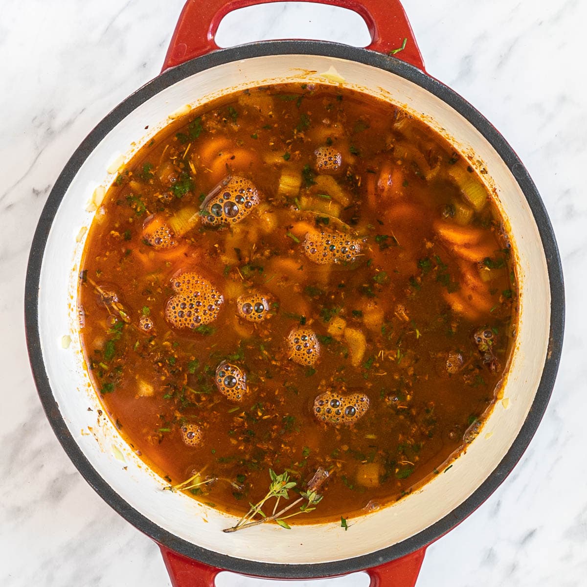 A red-white Dutch oven with dark brown stew and chopped onion, celery, and carrot slices.