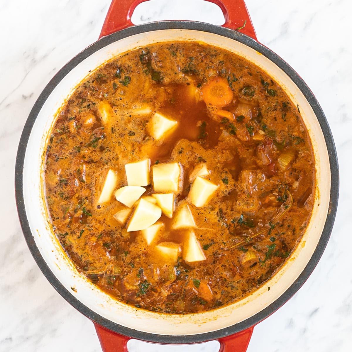 A red-white Dutch oven with dark brown stew and chopped onion, celery, potatoes, and carrot slices.