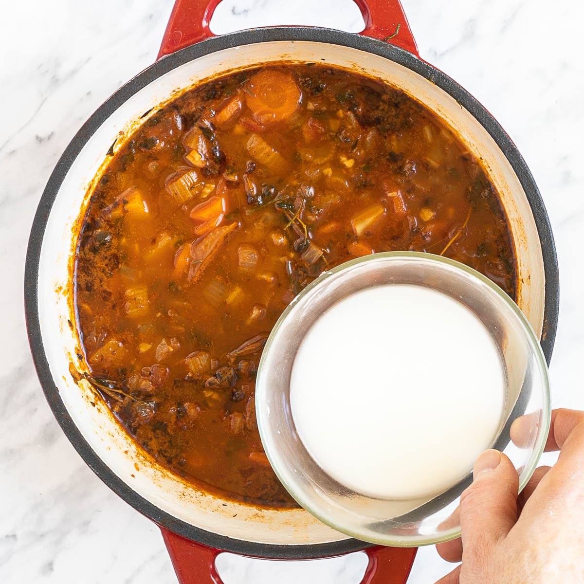 A red-white Dutch oven with dark brown stew and chopped onion, celery, potatoes, and carrot slices. A hand is holding a small bowl with a white liquid.