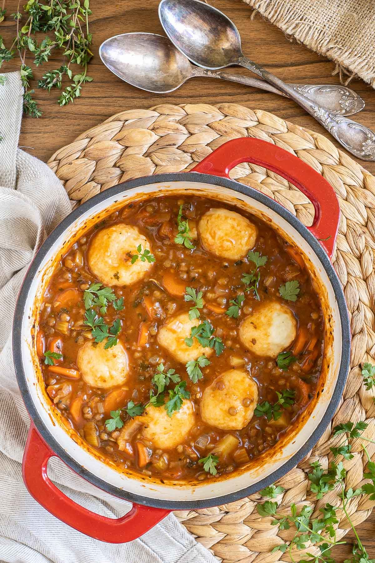 A red-white Dutch oven with thick brown stew, lentils, chopped veggies and a couple of dumplings.
