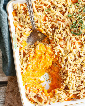 A white casserole dish from above showing orange mash topped with grated crumbles and thyme twigs. A serving spoon is dipped within.
