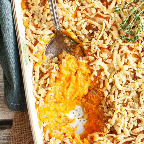 A white casserole dish from above showing orange mash topped with grated crumbles and thyme twigs. A serving spoon is dipped within.