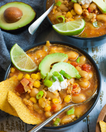 2 black bowls with orange liquid, beans, corn, chopped tomaotes, mince, avocado slices, chopped spring onion, lime slices, and tortilla chips. Half of avocado and more tortilla chips are scattered around