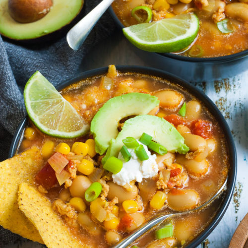 2 black bowls with orange liquid, beans, corn, chopped tomaotes, mince, avocado slices, chopped spring onion, lime slices, and tortilla chips. Half of avocado and more tortilla chips are scattered around