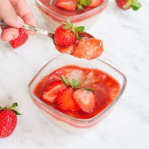 2 small glass bowls with white pudding with pearls topped with strawberry sauce and fresh strawberry slices. A hand is holding a spoon with a mouthful of the pudding.