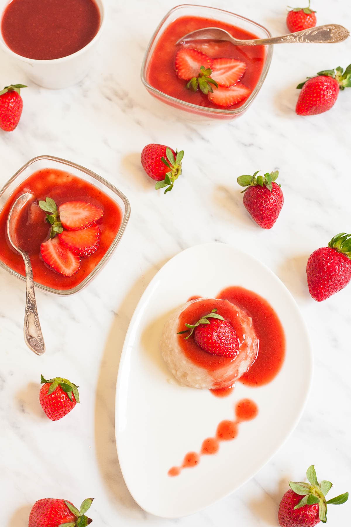 A small white plate with a heap of white pudding with pearls drizzled with strawberry sauce and topped with a half fresh strawberry. Other bowls of pudding and whole fresh strawberries are scattered around it.
