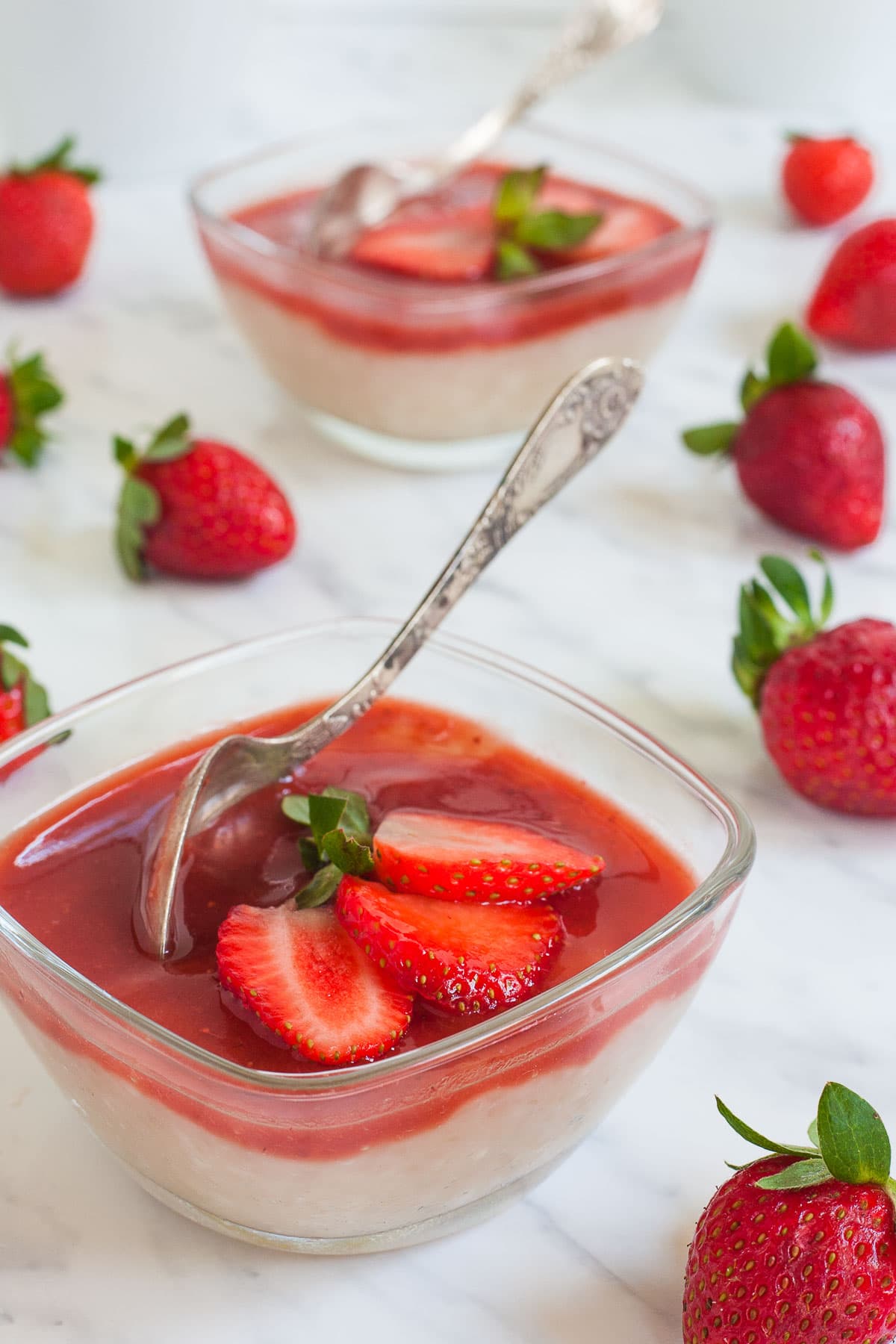 2 small glass bowls with white pudding with pearls topped with strawberry sauce and fresh strawberry slices. A spoon is placed inside.
