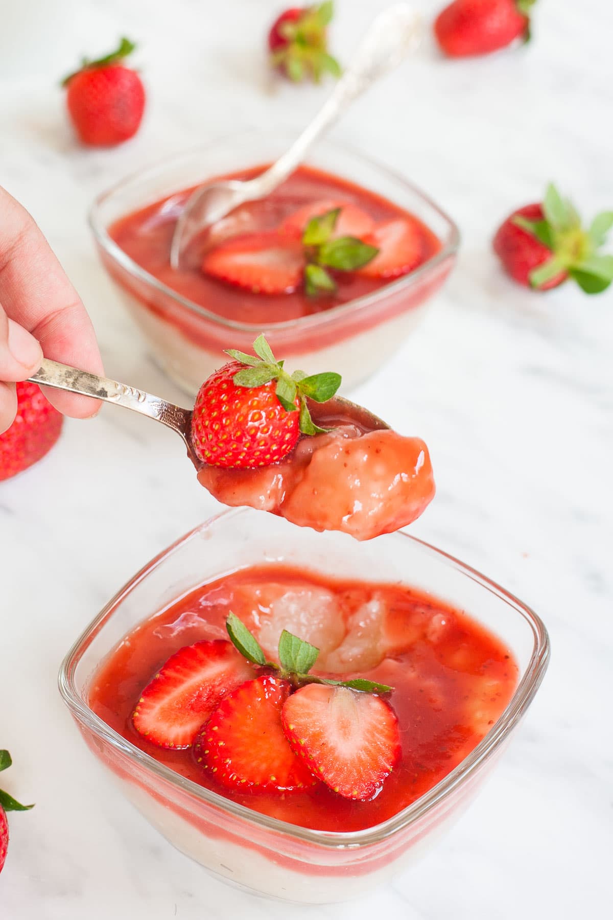 2 small glass bowls with white pudding with pearls topped with strawberry sauce and fresh strawberry slices. A hand is holding a spoon with a mouthful of the pudding.