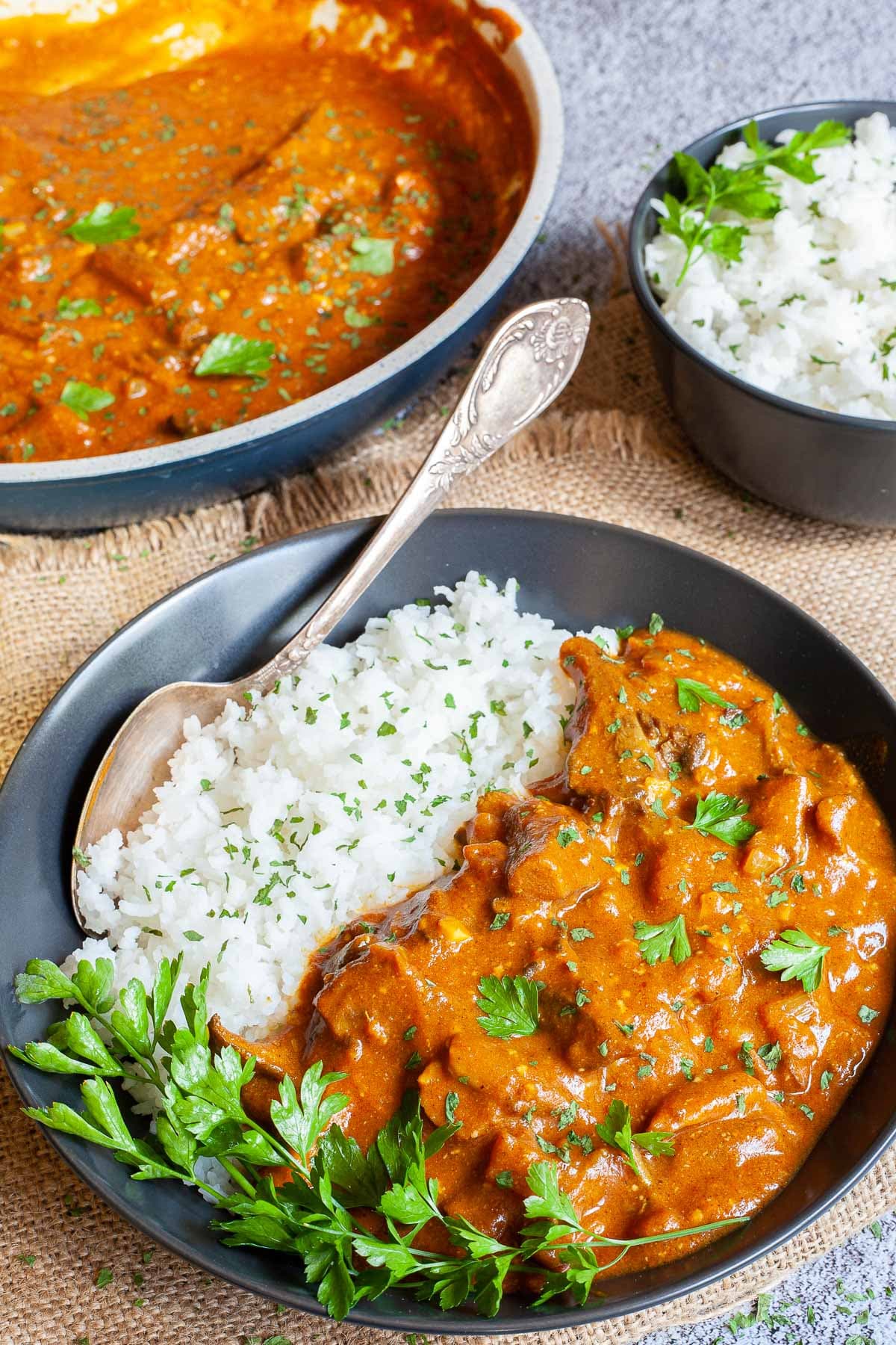 Large black plate with white rice and mushroom shreds in orange thick sauce sprinkled with chopped green herbs and a twig of parsley on top. Two spoons, a frying pan with sauce and a small bowl of leftover rice are placed next to it.