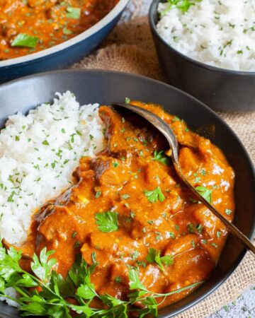 Large black plate with white rice and mushroom shreds in orange thick sauce sprinkled with chopped green herbs and a twig of parsley on top. Two spoons, a frying pan with sauce and a small bowl of leftover rice are placed next to it.