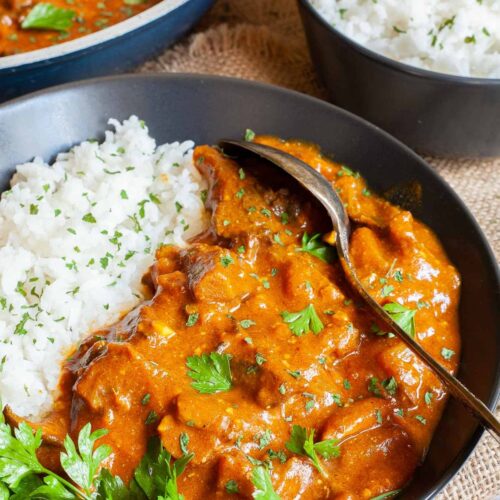 Large black plate with white rice and mushroom shreds in orange thick sauce sprinkled with chopped green herbs and a twig of parsley on top. Two spoons, a frying pan with sauce and a small bowl of leftover rice are placed next to it.