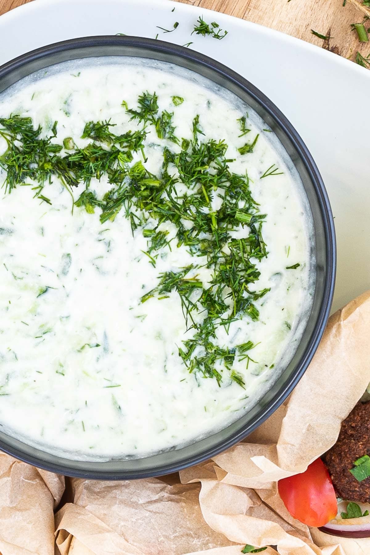 Black bowl with white green sauce topped with freshly chopped dill. The bowl is surrounded brown paper used as sandwich covers.