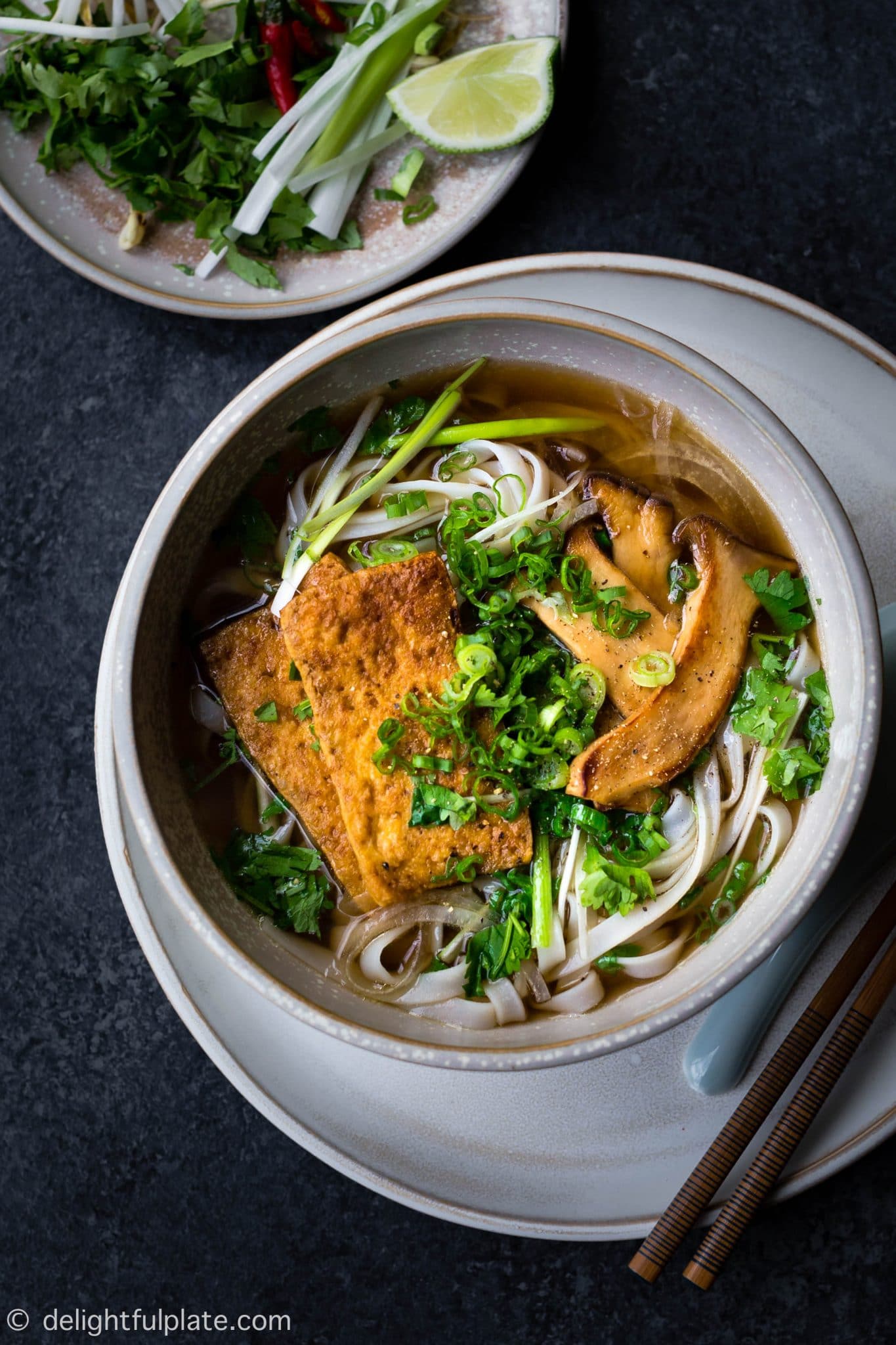 A dark brown soup served in a grey bowl with brown tofu slices, mushroom slices, noodles and lots of green leaves