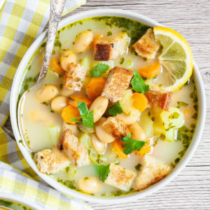 Light yellow soup served in a white bowl with a spoon full of large white beans, croutons, carrot slices, diced potatoes and freshly chopped herbs.