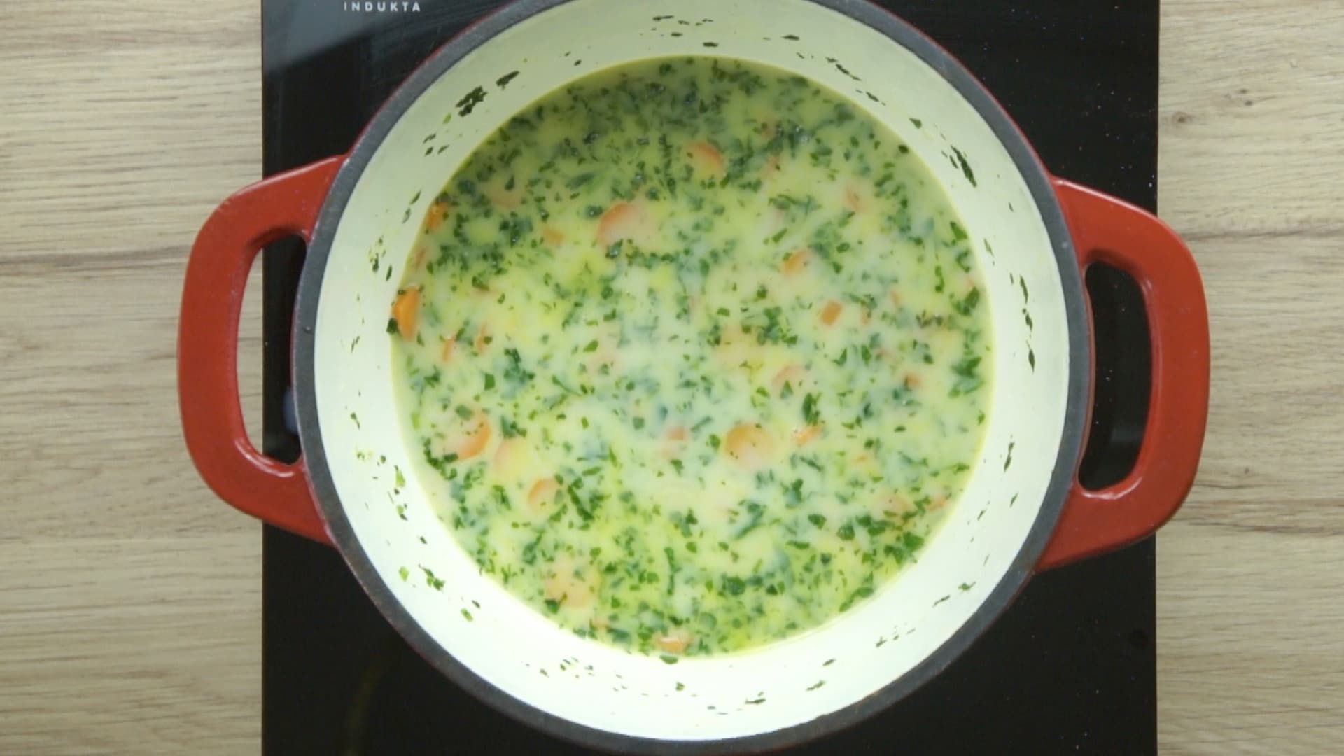 Carrot slices, parsnip slices, and freshly chopped green herbs are in a light yellow soup in a red enameled Dutch oven.